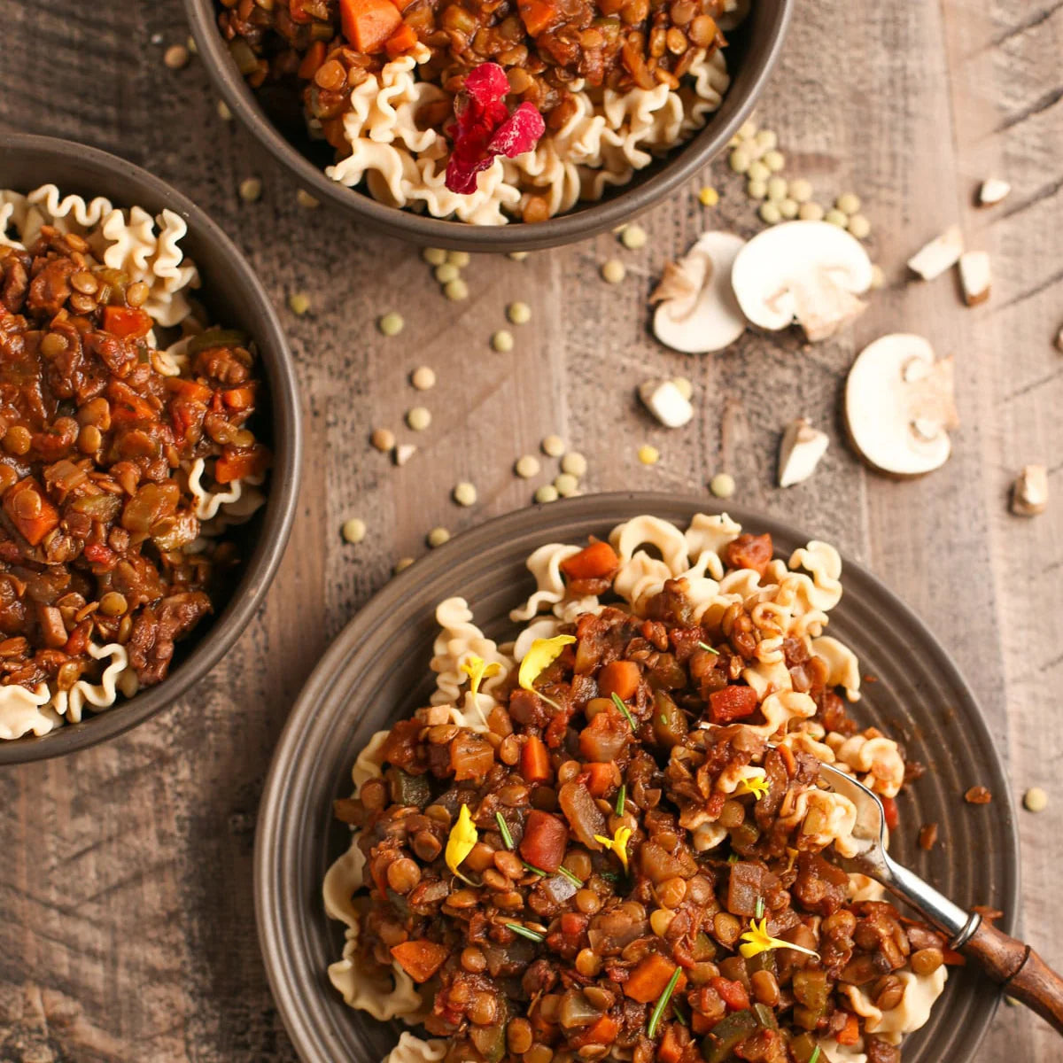 Three bowls of pasta with a hearty lentil and carrot stew, arranged on a rustic wooden table with scattered lentils and mushrooms.