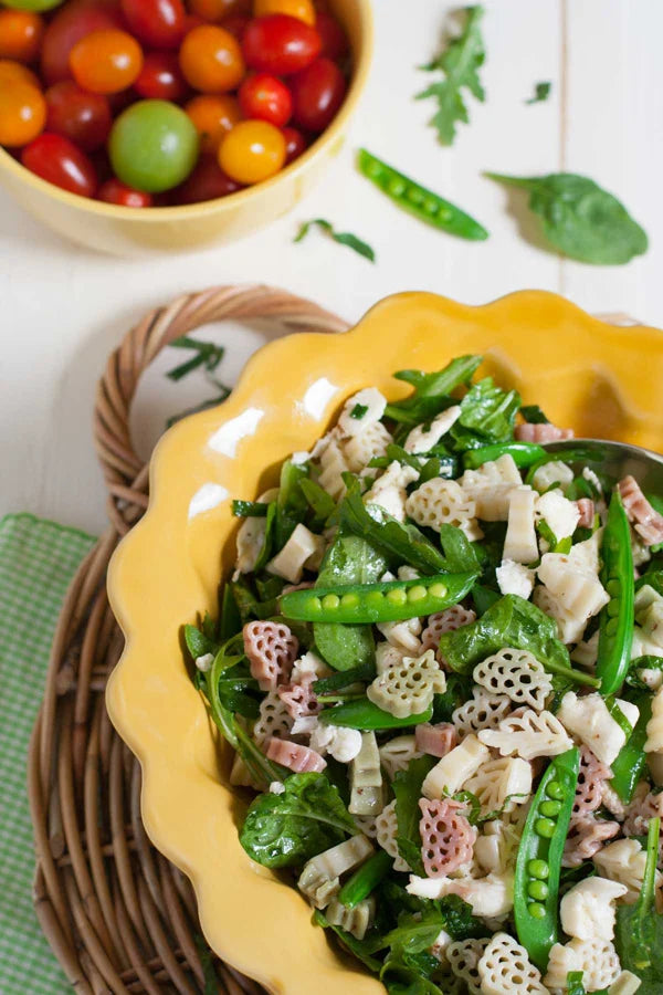 A close-up of a fresh salad in a yellow scalloped bowl, featuring green peas, spinach, and patterned pasta, with a bowl of cherry tomatoes in the background on a white wooden surface.