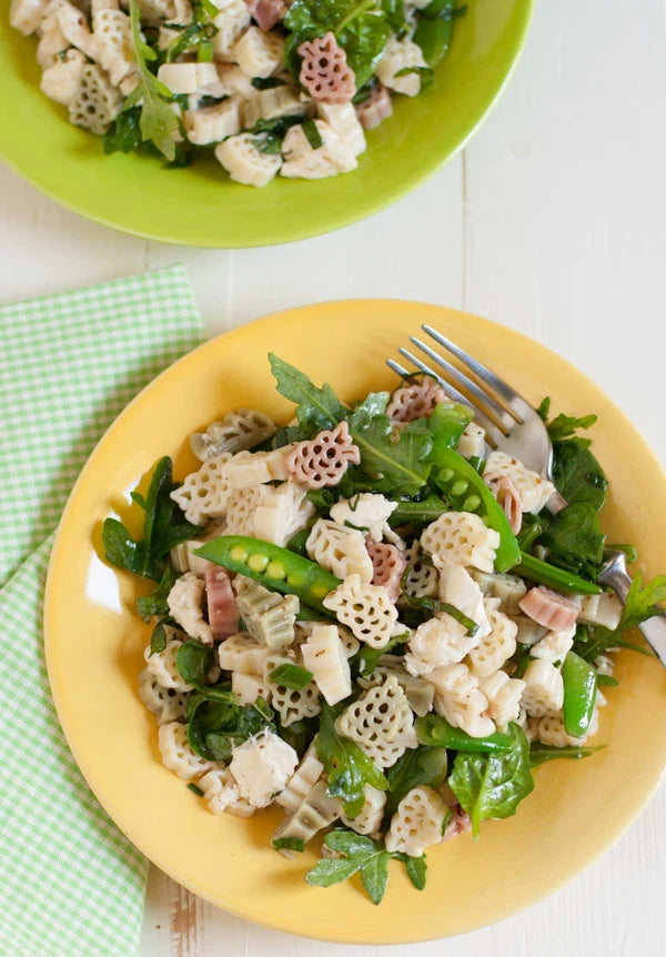 A close-up overhead shot of a pasta salad served on a yellow plate, featuring decorative pasta, green beans, spinach, and pieces of chicken, with a fork resting on the plate. A second green plate with more salad is visible in the upper background on a white wooden surface next to a green checkered napkin.