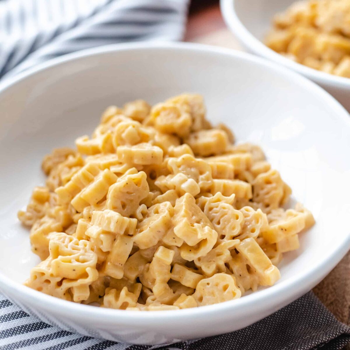 A close-up of a white bowl filled with creamy macaroni pasta, with a second bowl blurred in the background on a wooden surface.