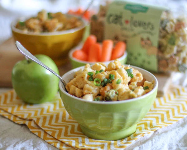 A bowl of creamy chicken and vegetable pasta salad garnished with chopped green onions, set on a yellow and white chevron napkin. A green apple, baby carrots, and a bag of Cat Lovers pasta are visible in the softly blurred background.