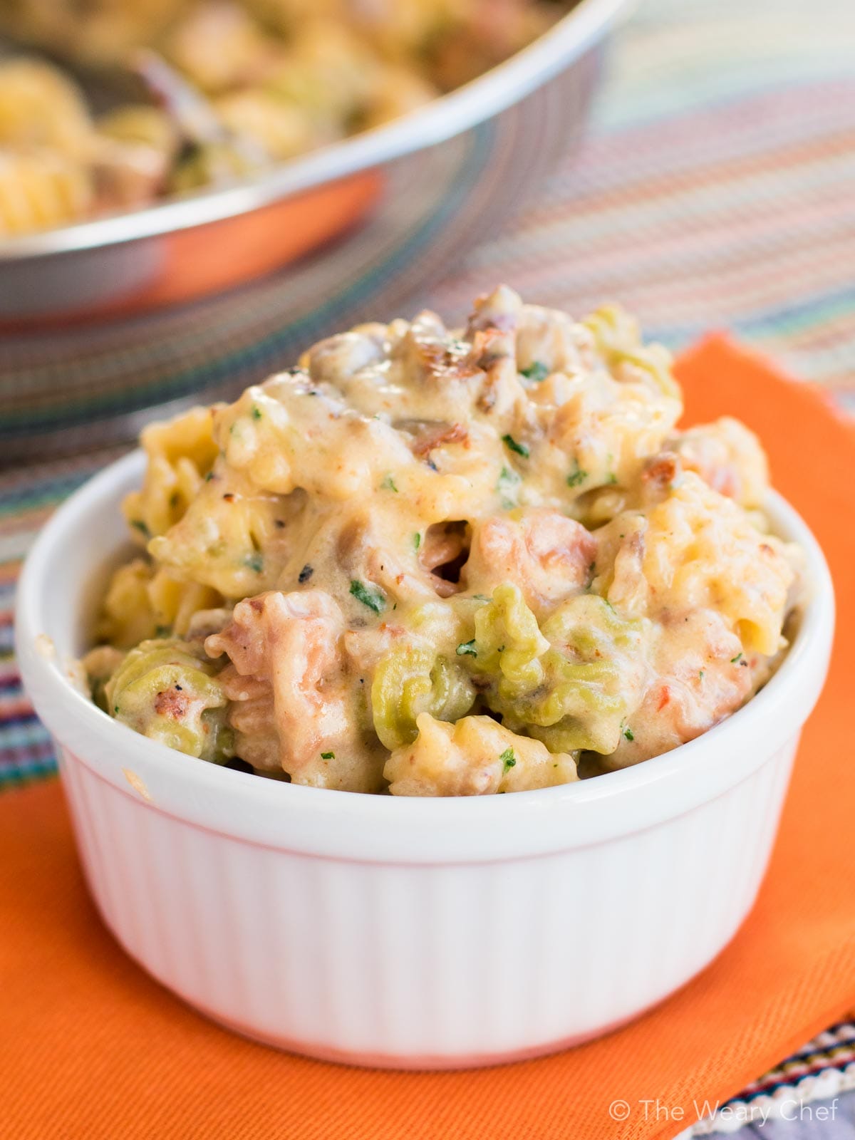 A close-up shot of a creamy pasta salad with chicken and broccoli served in a small white ramekin, with a larger bowl blurred in the background on a striped tablecloth.