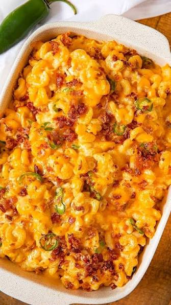 A close-up overhead shot of a baked macaroni cheese casserole in a white rectangular baking dish, topped with melted cheese, crispy browned bits, and sliced green peppers. A whole green jalapeño is visible in the top-left corner on a white cloth, with a wooden surface underneath.