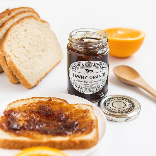 Jar of Tawny Orange jam with a slice of toast spread with jam, surrounded by bread slices and an orange.