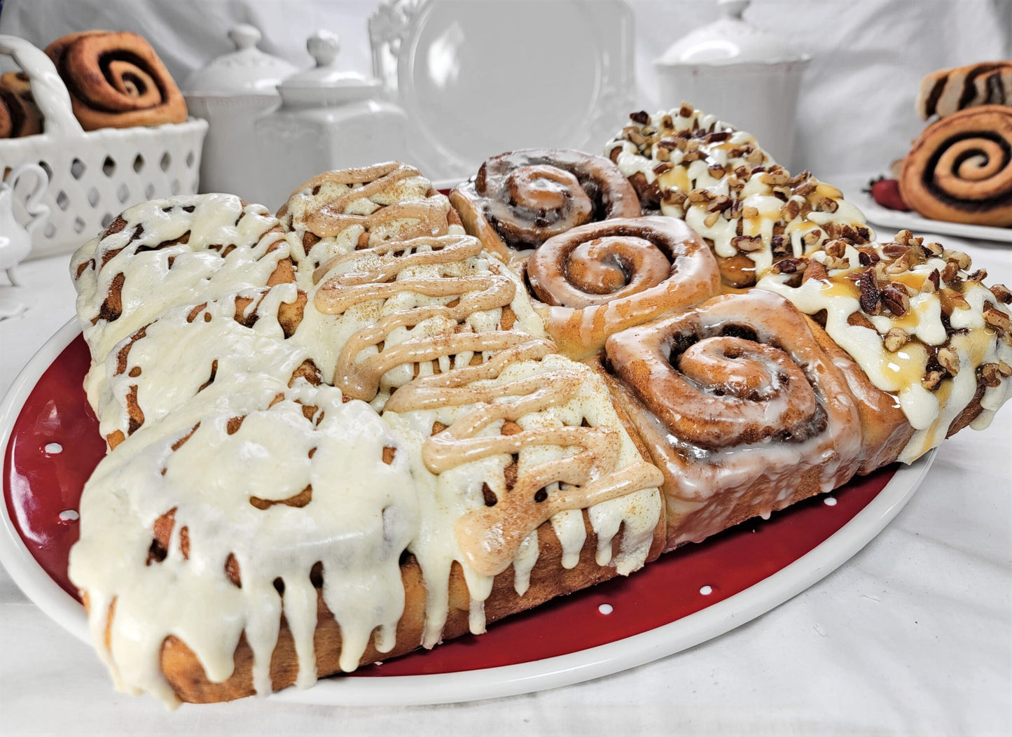 A large assortment of baked cinnamon rolls and sweet rolls arranged on a red and white oval platter, with white icing, drizzles, and chopped nuts on top. The pastries are photographed in a styled kitchen or bakery setting with white ceramic dishes and baking items blurred in the background.