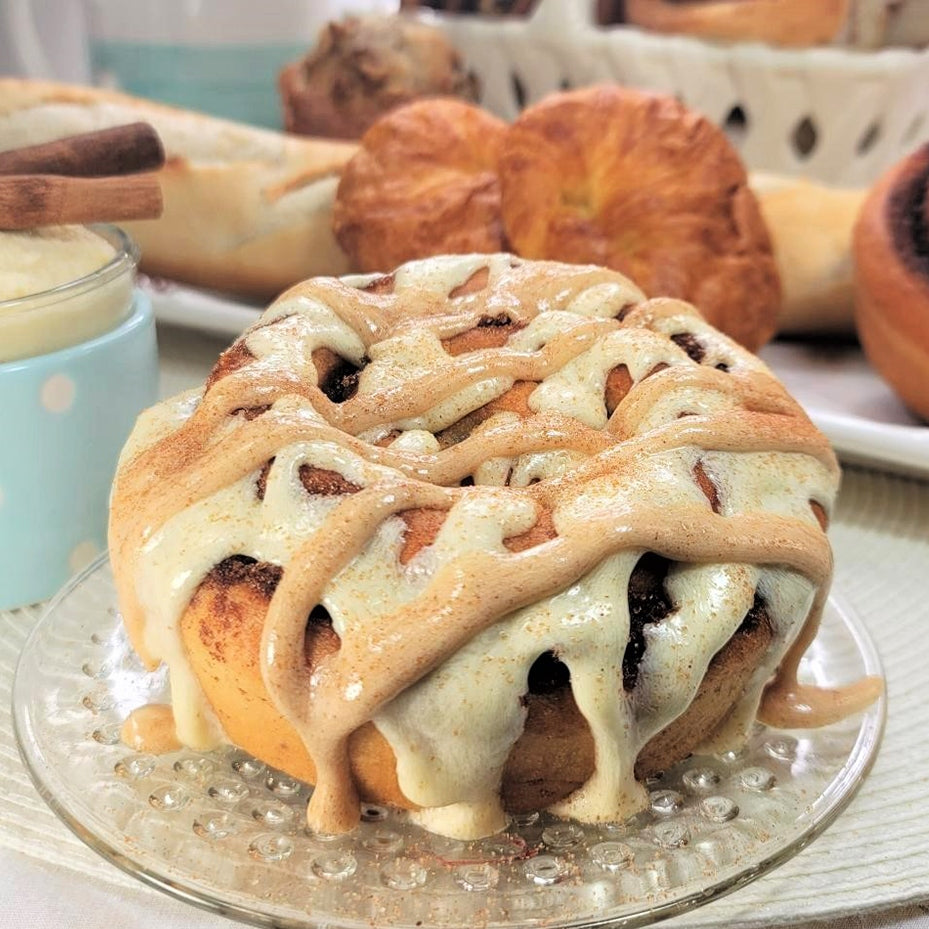 A close-up of a cinnamon roll on a clear glass plate, topped with a light beige icing drizzle and sprinkled with cinnamon. Other pastries are blurred in the background on a table.
