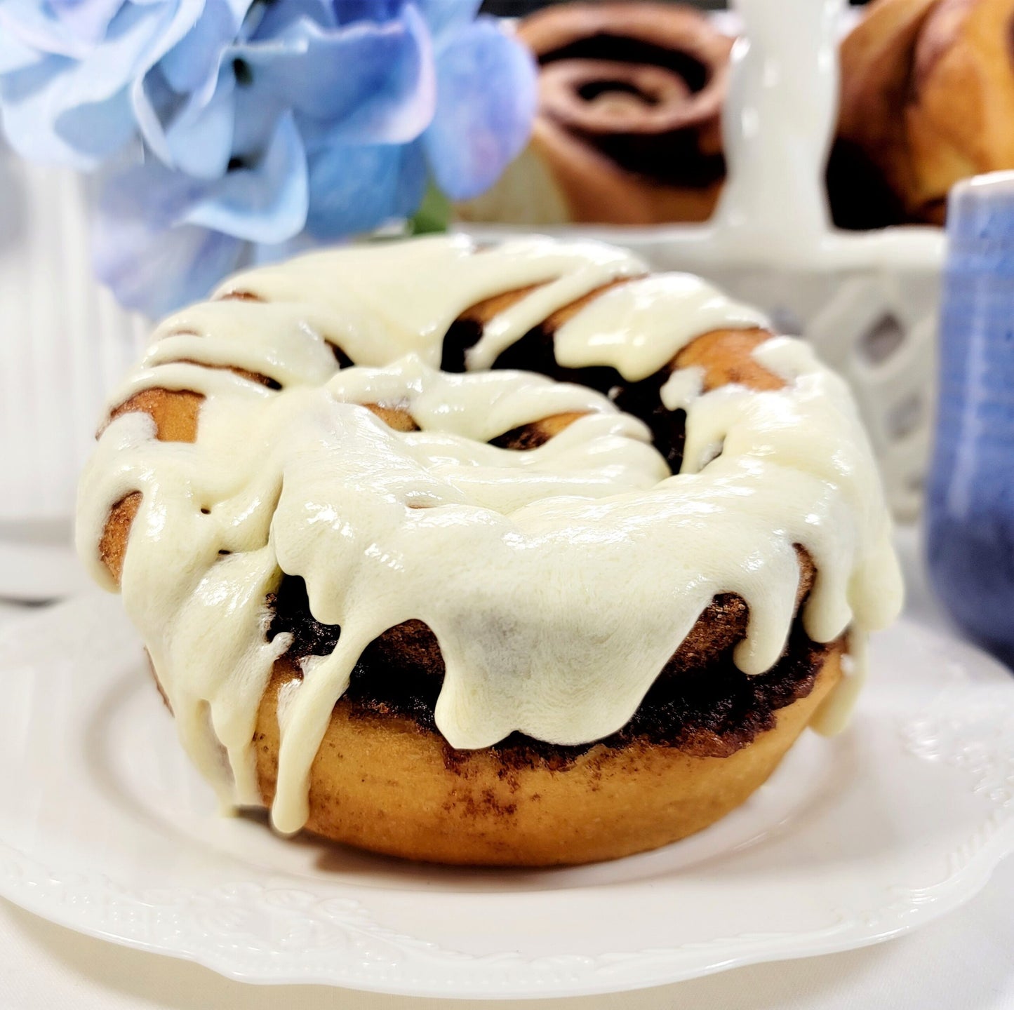 A close-up of a cinnamon roll or cinnamon bun topped with thick white icing, served on a white plate with a softly blurred kitchen or bakery background.