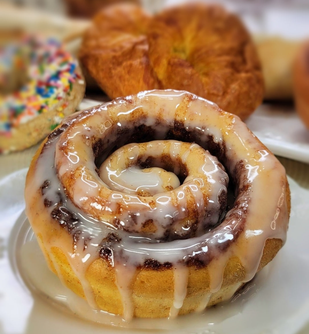 A close-up of a glazed cinnamon roll on a white plate, with other pastries blurred in the background.