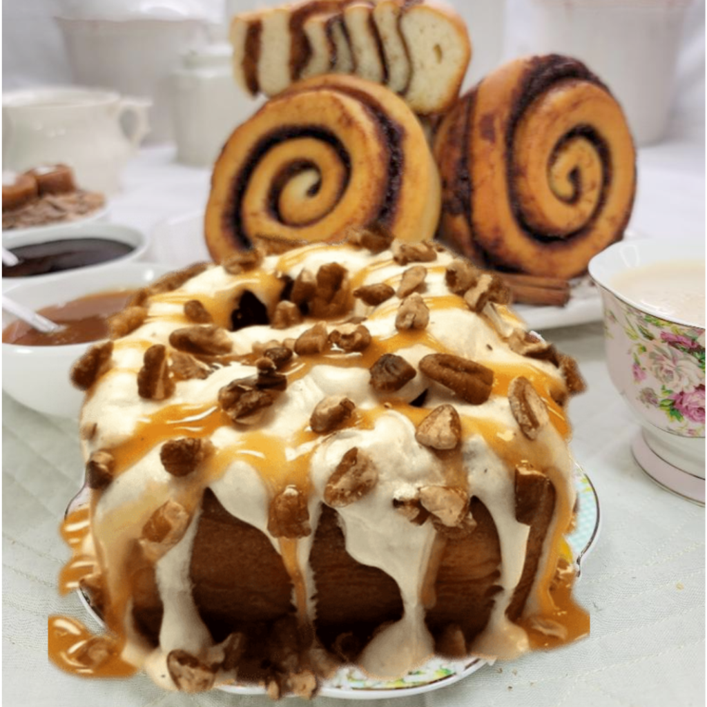 A large round cinnamon roll cake or bread covered with caramel sauce and chopped nuts, served on a plate in a bakery setting with sliced cinnamon rolls and tea cups in the background.