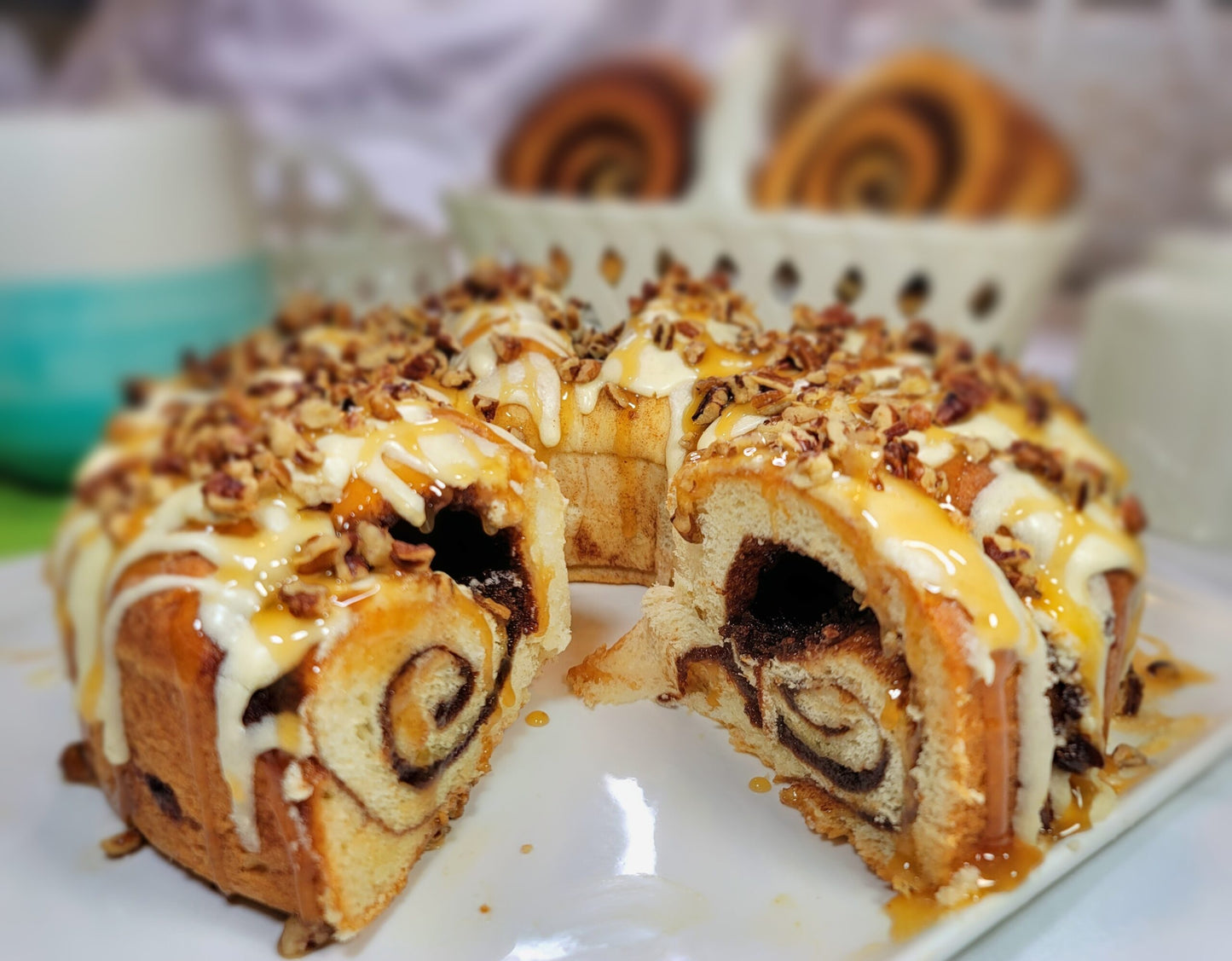 A close-up of a sliced cinnamon roll cake on a white plate, topped with white icing and chopped nuts, with more cinnamon rolls blurred in the background.
