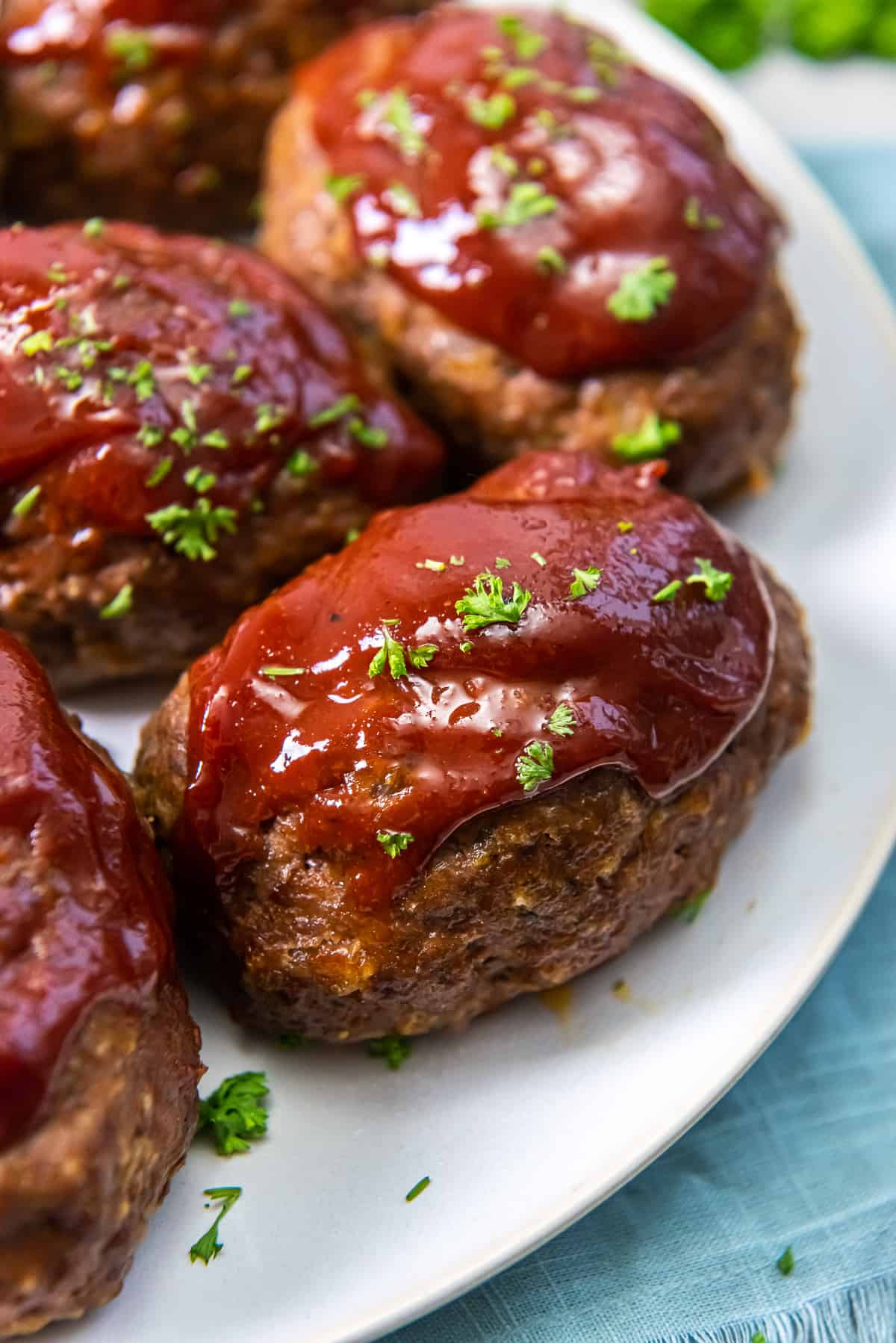 Close-up of several meatloaf patties topped with glossy red sauce and chopped green parsley, served on a white plate over a blue cloth background.