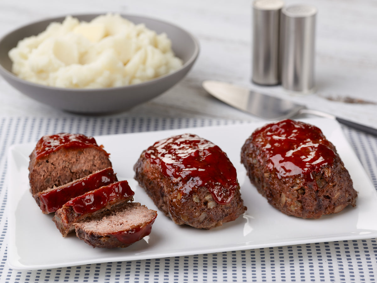 Three glazed meatloaves on a white rectangular plate, with one slice cut and placed beside them. A bowl of mashed potatoes, a knife, and metal salt and pepper shakers are visible in the background on a light wood surface with a blue-and-white striped napkin.