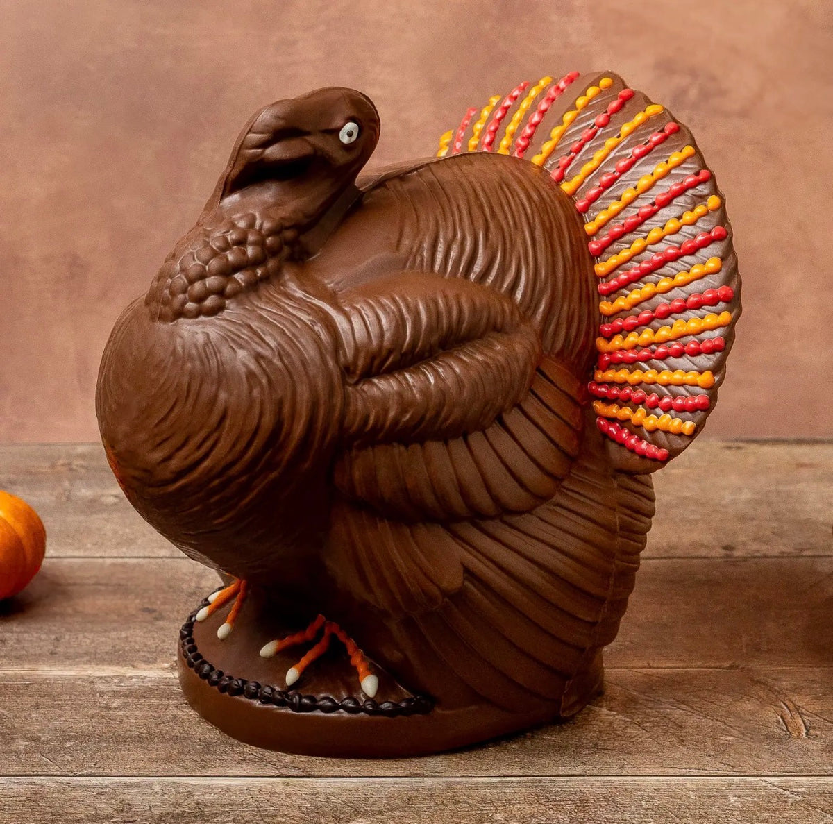 A decorative Chocolate turkey centerpiece or ornament in dark brown with a bright multicolor tail feathers pattern of red, orange, yellow, and gray, displayed on a rustic wooden surface against a warm brown background.