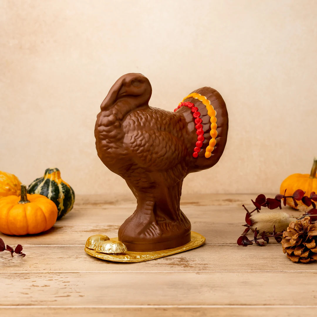 A chocolate turkey figurine with a gold foil base, decorated with red and yellow candy-like stripes on its tail, displayed on a wooden surface with small pumpkins and autumn decor in the background.