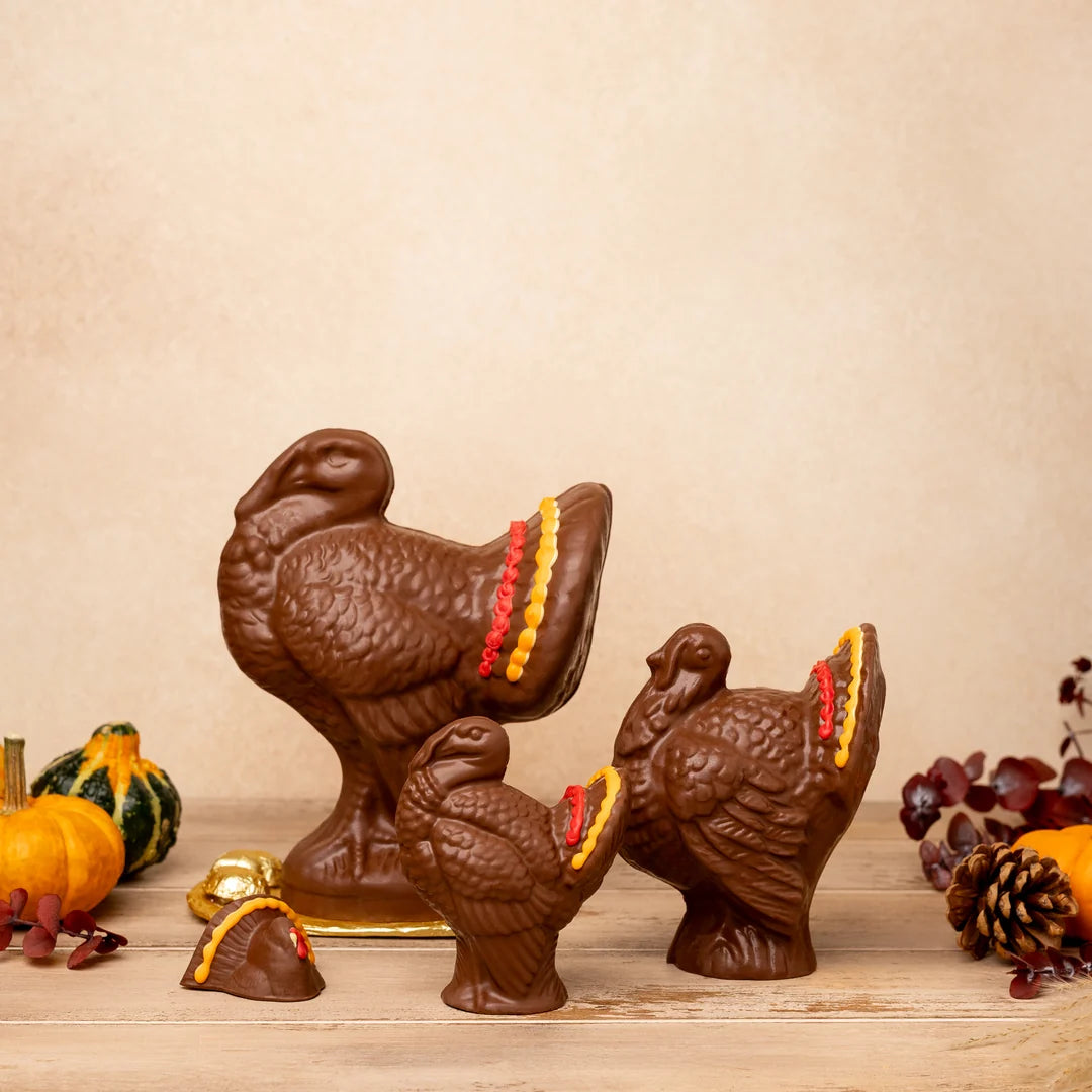 Three brown turkey-shaped chocolate ornaments displayed on a wooden surface against a warm beige background, with small decorative pumpkins and dried leaves around them.