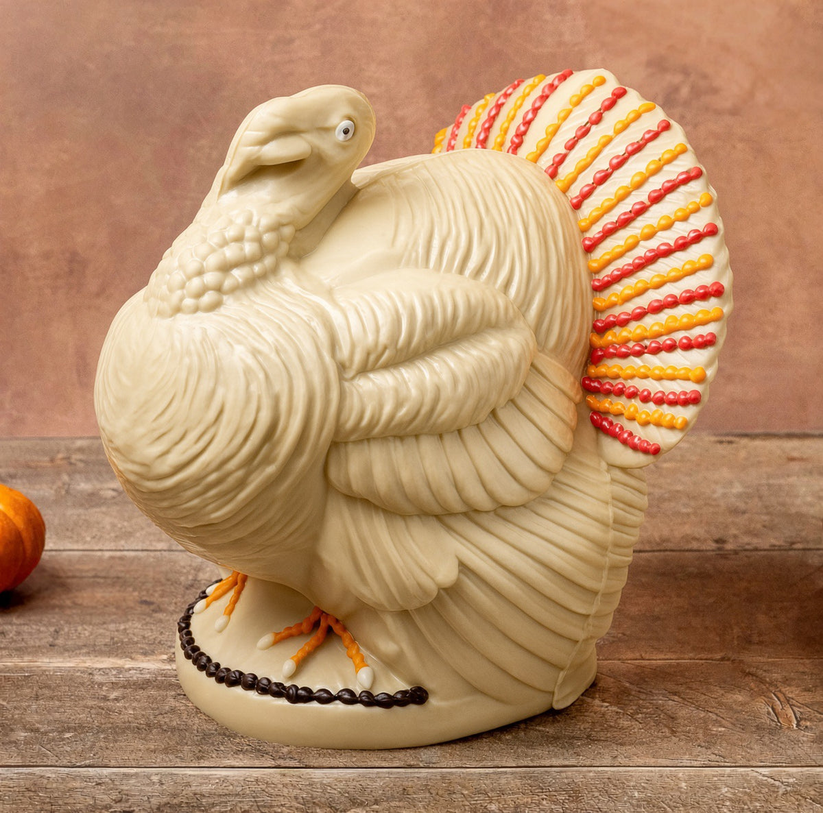 A white chocolate Thanksgiving turkey centerpiece in cream with a striped red, orange, and yellow tail, standing on a wooden surface against a brown background.
