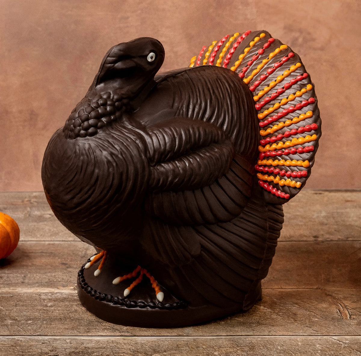 A decorative chocolate turkey centerpiece in dark brown with a vibrant fanned tail in red, orange, yellow, and gray, displayed on a rustic wooden surface against a warm brown background.