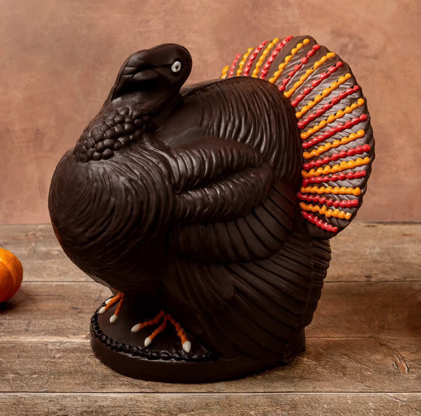 A decorative chocolate turkey centerpiece in dark brown with a vibrant fanned tail in red, orange, yellow, and gray, displayed on a rustic wooden surface against a warm brown background.