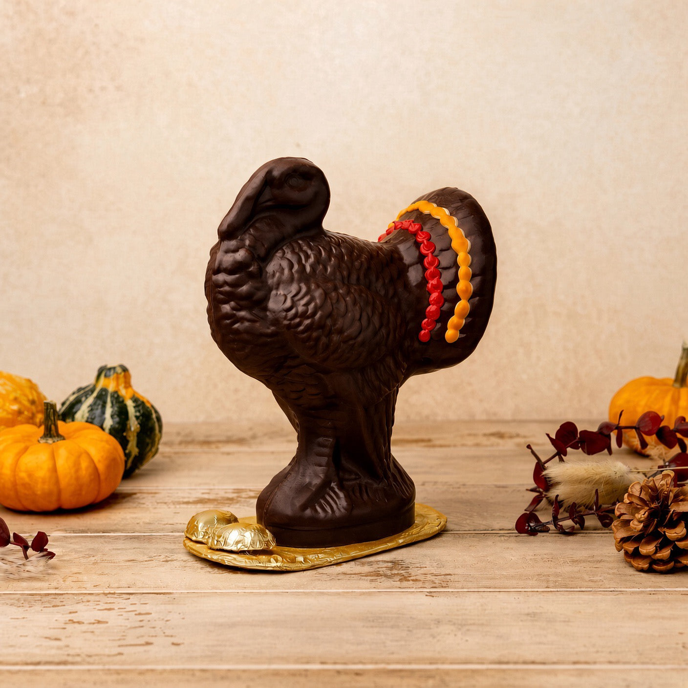 A dark brown chocolate turkey figurine with a gold base, decorated with red, orange, and yellow candy accents on its tail feathers, displayed on a rustic wooden surface with autumn decorations.