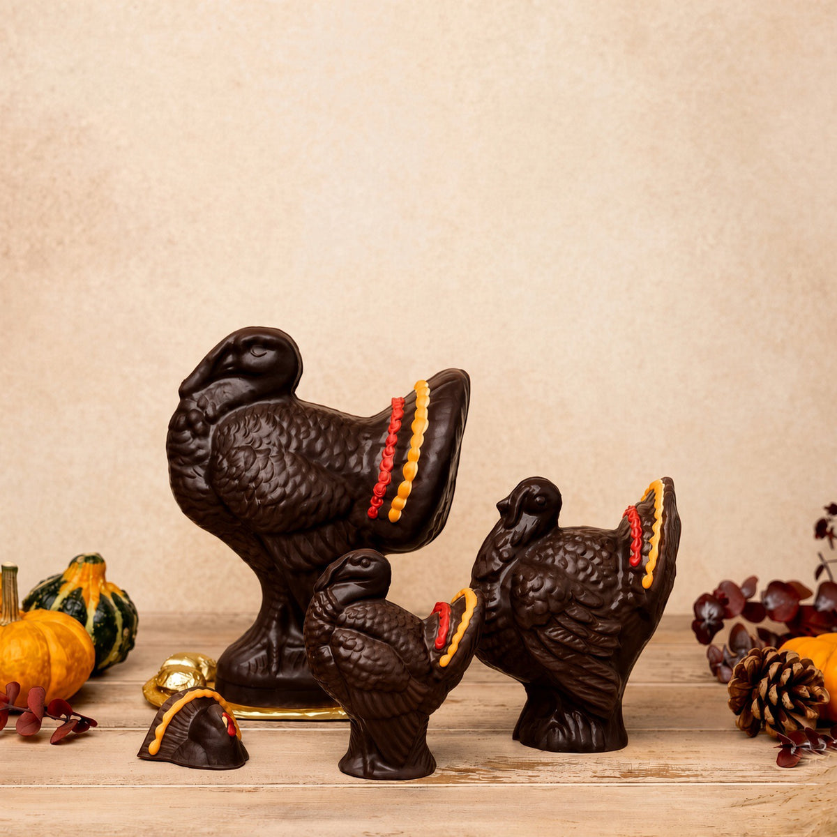 Three dark brown turkey figurines with red, orange, and yellow feather accents arranged on a wooden surface against a beige background, with small decorative pumpkins and pinecones around them.