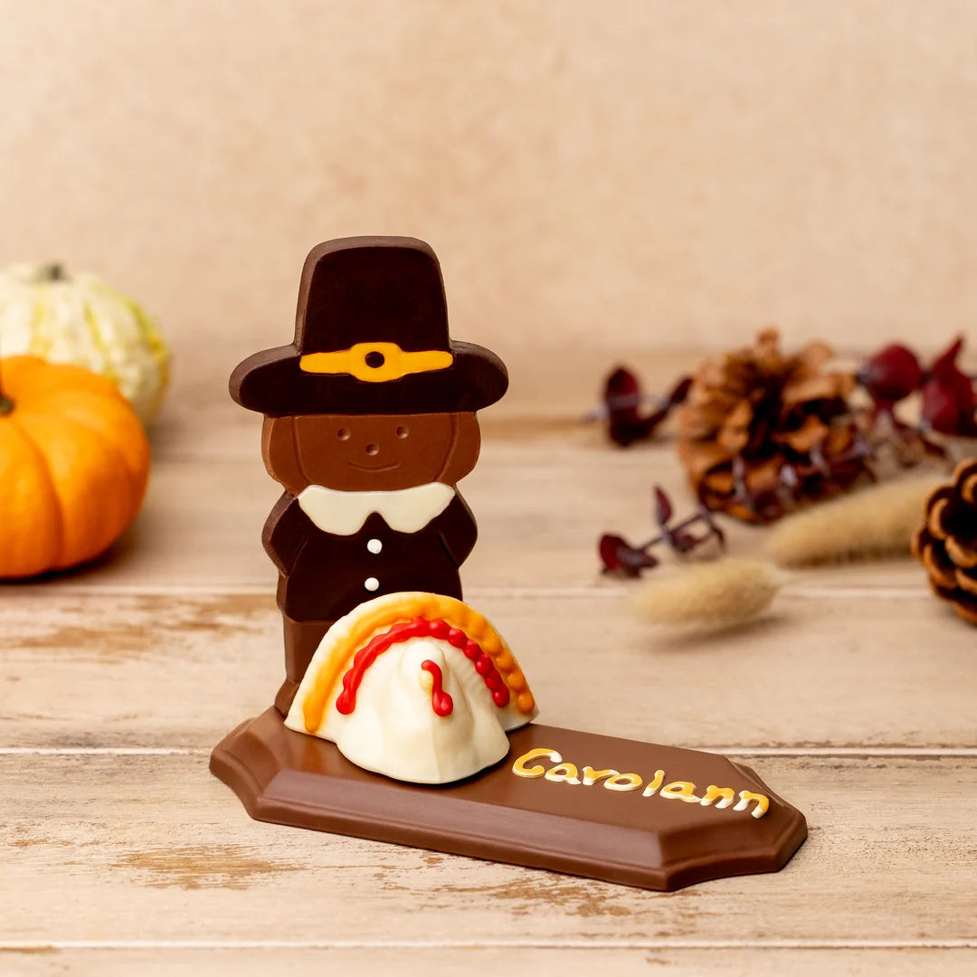 A personalized chocolate haped like a Thanksgiving pilgrim standing beside a white and orange pie, mounted on a brown base engraved with the name 'Carolann'. The scene is styled on a wooden surface with autumn props including pumpkins, pinecones, and dried grass in the background.