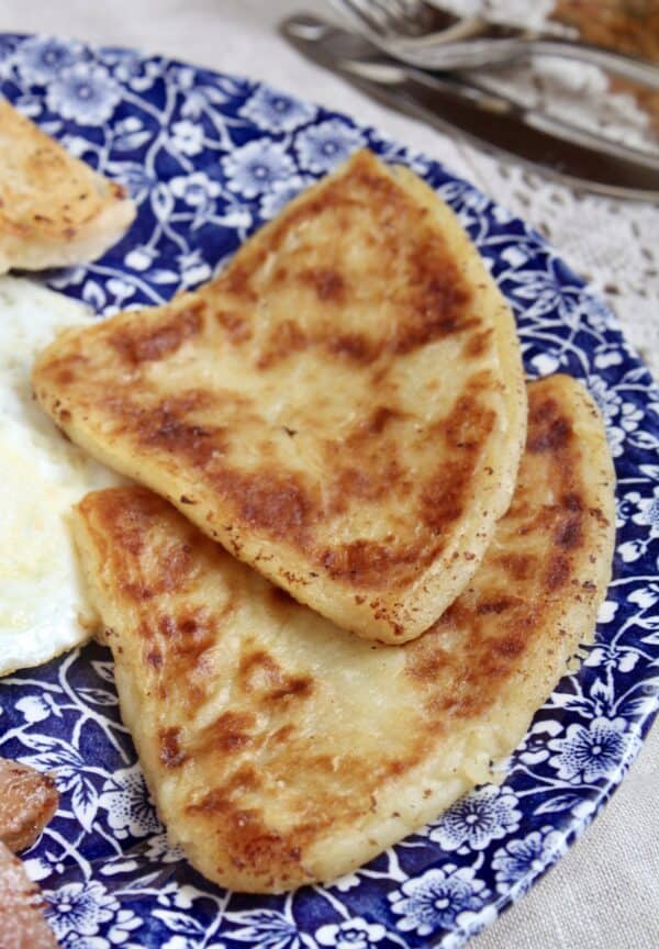 Three Irish potato bread cakes served on a blue and white floral patterned plate, with a fried egg and silver cutlery partially visible in the background.