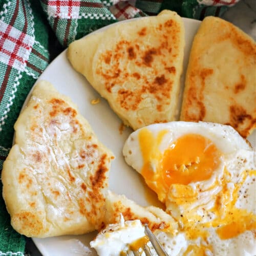 A close-up of a breakfast plate featuring golden-brown baked Irish potato bread cakes with a sunny-side-up egg that is broken open, showing a bright orange yolk, on a white plate with a fork.
