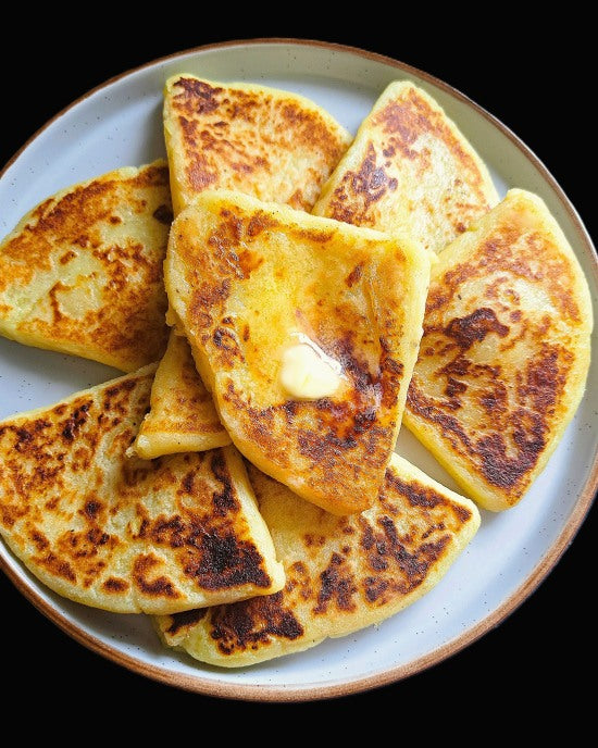 A top-down view of golden-brown Irish potato bread cakes on a speckled white plate with a brown rim, topped with a melting pat of butter against a black background.