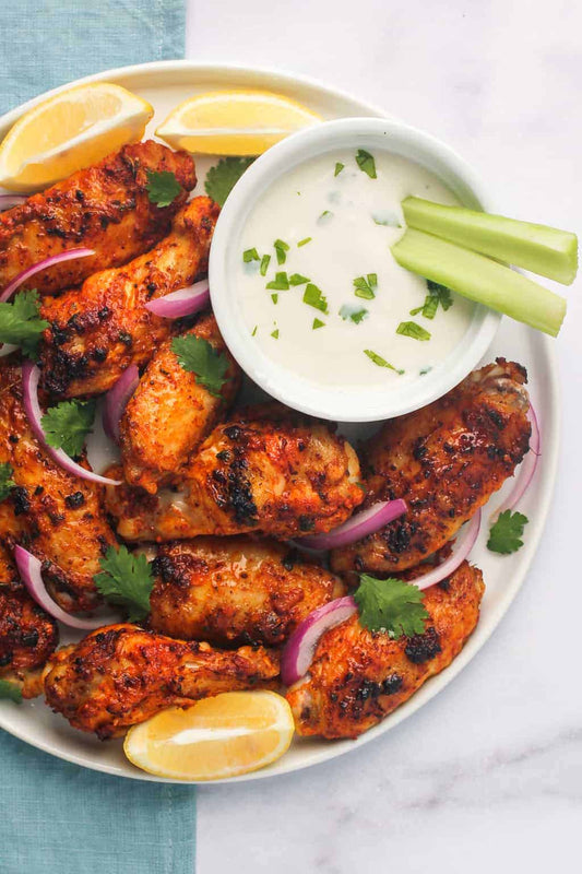 Grilled tandori chicken wings served on a white plate with a side of creamy white dipping sauce, garnished with red onion, cilantro, lemon wedges, and celery sticks, set on a light blue napkin over a white marble surface.