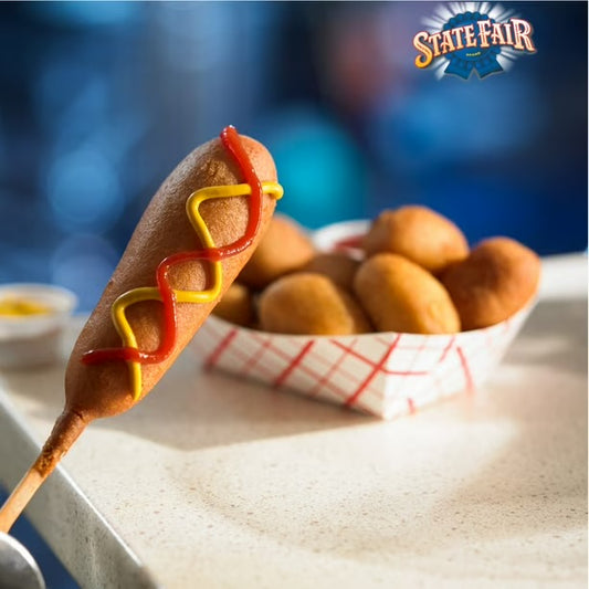 A hot dog corn dog on a wooden stick, topped with red ketchup and yellow mustard, with a basket of fried dough balls in the background. A 'State Fair' logo is visible in the top right corner.