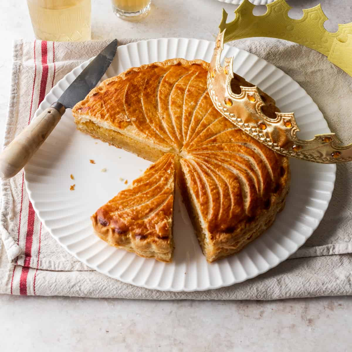 A golden-brown baked pastry pie with a decorative leaf pattern on top, served on a white plate with a slice cut out. A gold crown decoration and a knife are placed beside the plate on a striped napkin.