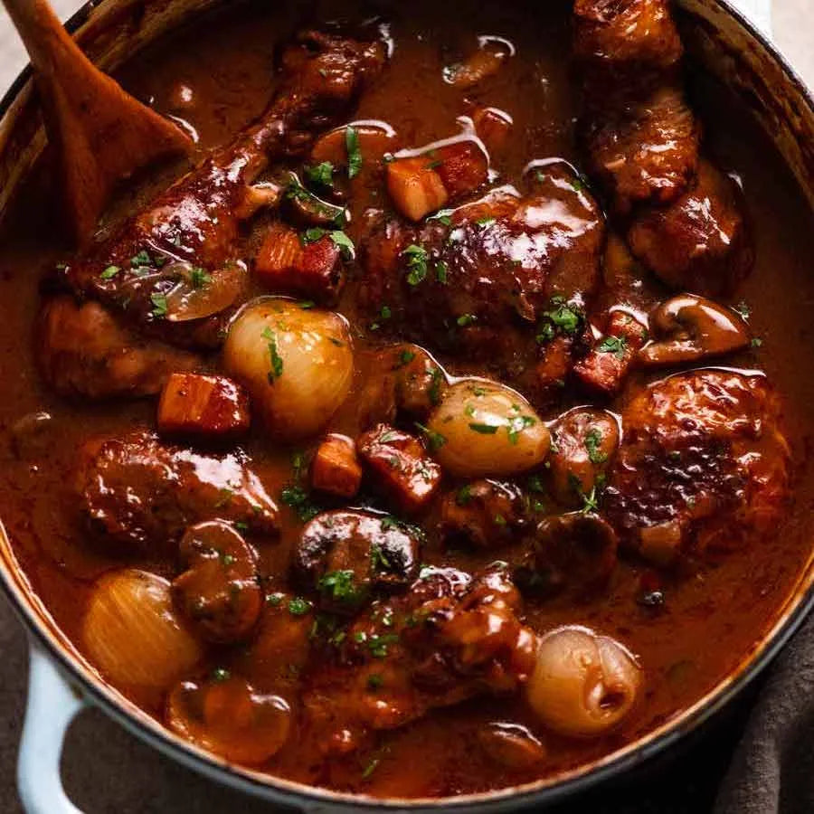 A close-up overhead shot of a rich, glossy chicken and mushroom stew in a dark brown sauce, served in a white enameled cast iron pot with a wooden spoon.
