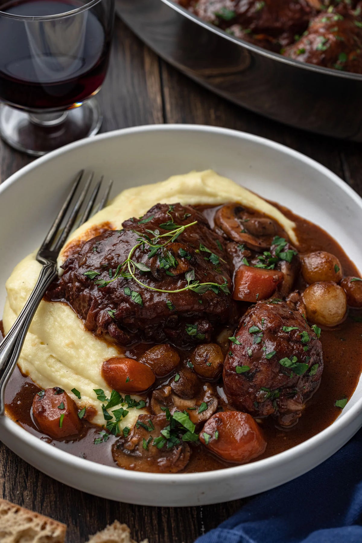 A plated beef stew dish with mashed potatoes, mushrooms, carrots, and herbs on a dark wooden table, with a glass of red wine and a pan in the background.