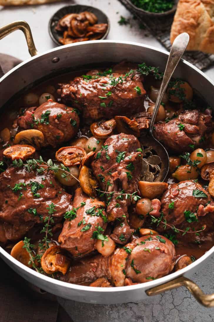 A close-up overhead shot of a large metal pot filled with braised chicken pieces in a rich brown sauce, mixed with sliced mushrooms, pearl onions, and chopped green herbs, with a metal serving spoon resting inside.