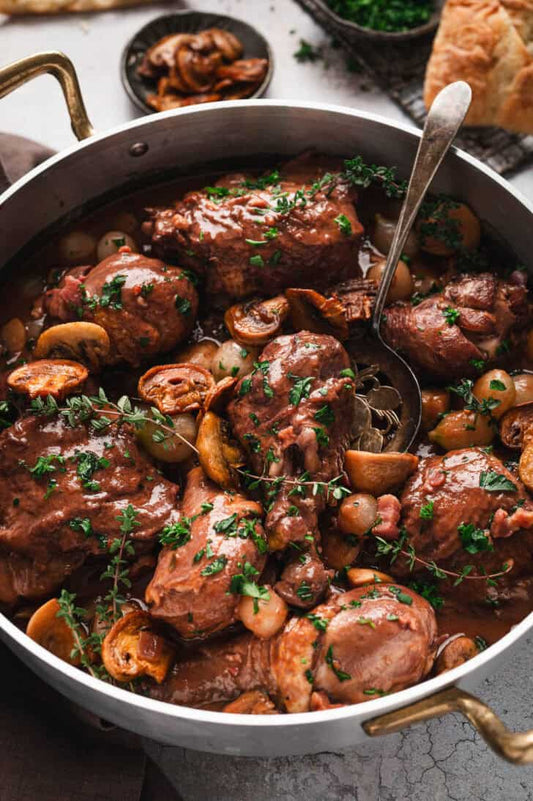 A close-up overhead shot of a large metal pot filled with braised chicken pieces in a rich brown sauce, mixed with sliced mushrooms, pearl onions, and chopped green herbs, with a metal serving spoon resting inside.