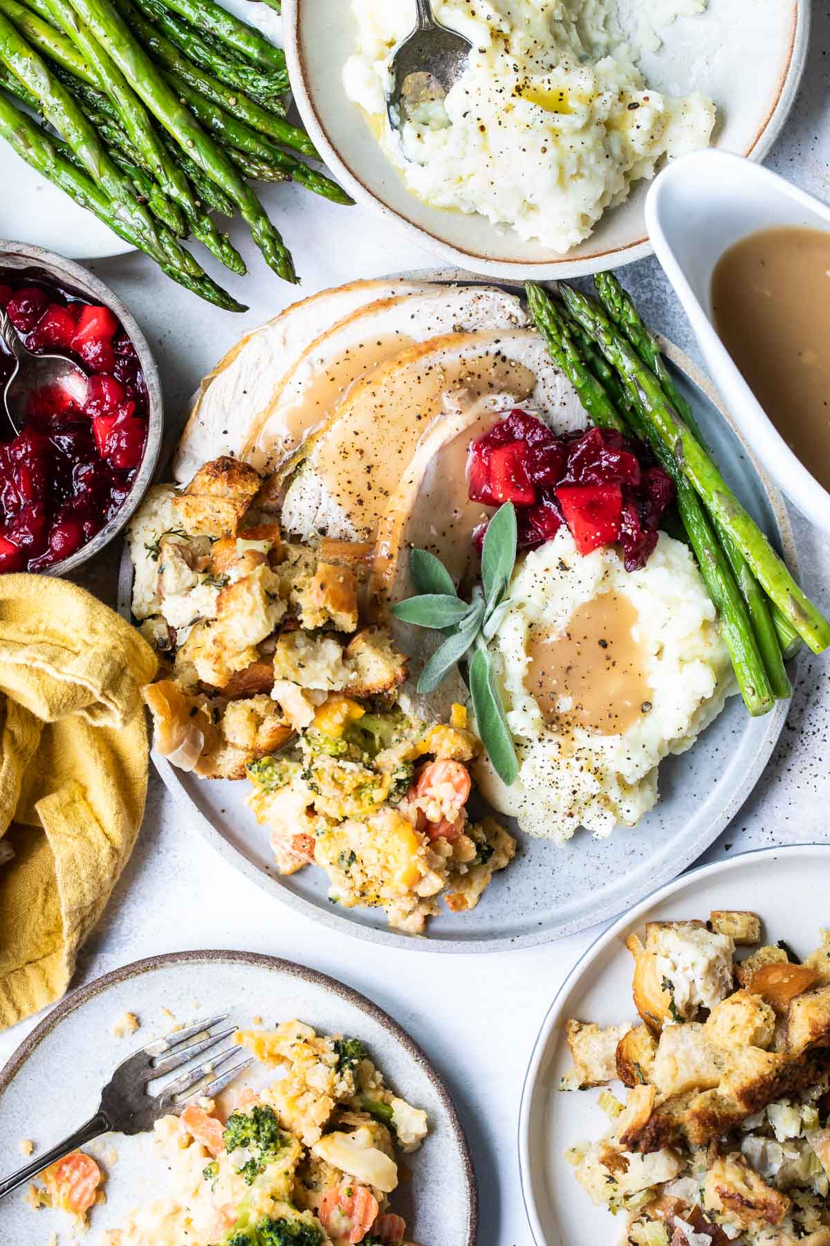 Overhead view of a Thanksgiving meal spread with plates of mashed potatoes, stuffing, cranberry sauce, roasted turkey, asparagus, gravy, and mashed potatoes on a light surface.