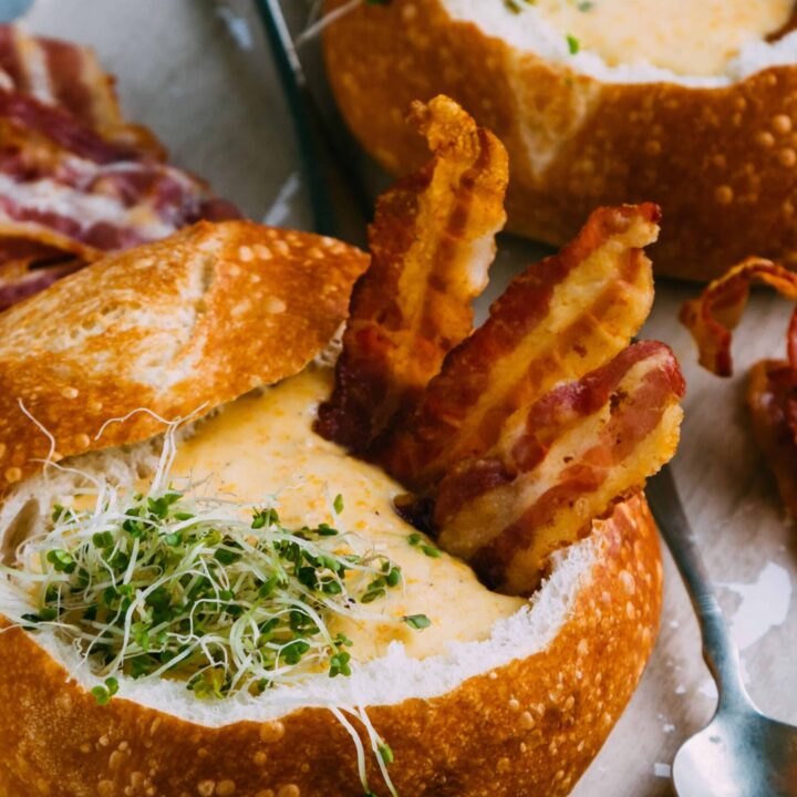 Soup & bread pairings, chili kits, bread-only four-packs, petite boules, and bulk cases — all ready for your coziest fall meals. A close-up of a gourmet food dish: a hollowed-out crusty bread bowl filled with melted cheese and topped with crispy bacon strips and green sprouts, served on a plate.