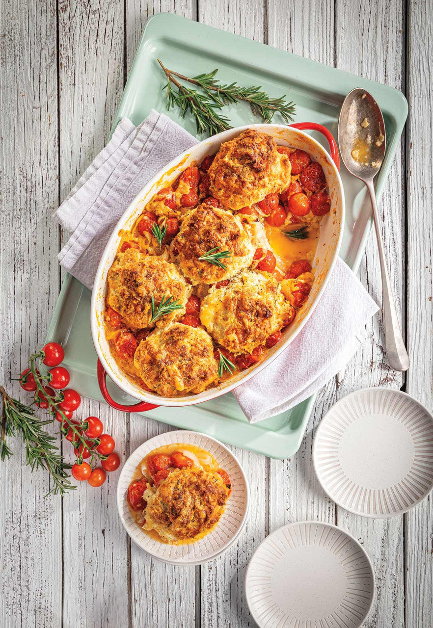 A baked dish of golden-brown bread or cheese cakes with tomato sauce and herbs, served in an oval white casserole dish on a mint green tray, with a smaller plated portion in front on a rustic white wood table.