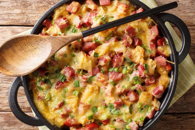 A close-up overhead shot of a baked cornbread casserole with diced sausage and chopped green herbs, served in a black cast-iron skillet with a wooden spoon resting across the top on a rustic wooden table.