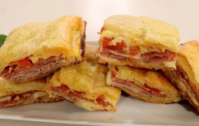 Close-up of several golden-brown baked stuffed pastries or sandwiches cut into pieces on a white plate, showing melted cheese and red filling.
