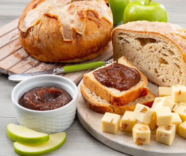 A styled food scene showing artisanal bread, sliced bread spread with dark jam, cubed cheese, and green apple slices on a light wood surface.