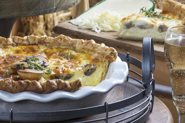 A baked mushroom and cheese quiche in a flaky pastry crust, served in a white ceramic pie dish on a black metal serving tray. A glass of sparkling beverage and another quiche on a cutting board are visible in the background.