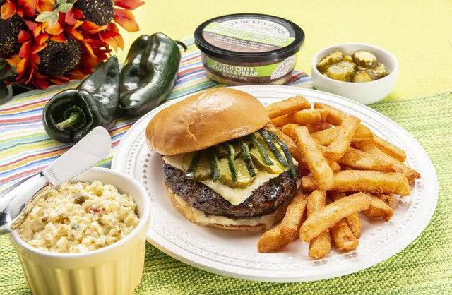 A plated burger with pickled jalapeño slices, cheese, and fries on a white plate, served with coleslaw, a side of sliced pickles, and a container of gourmet sauce on a bright green table setting.