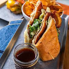 A close-up food photo of a large sandwich on a metal tray, served with potato chips, a small glass jar of dark sauce, and a blue napkin.
