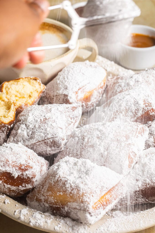 Powdered sugar being sifted over golden beignets on a plate beside coffee
