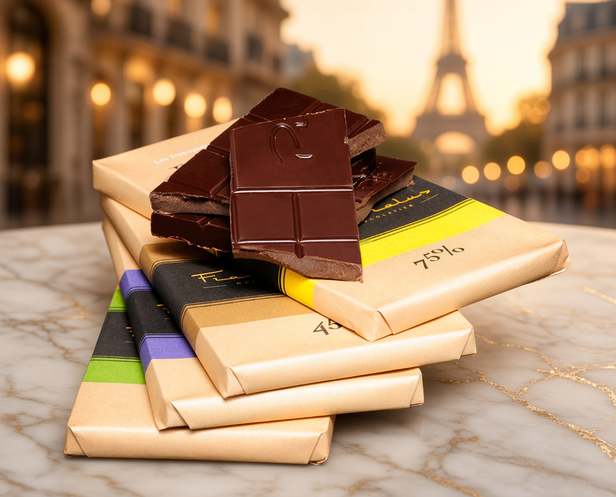 A stack of chocolate bars in kraft paper packaging with one broken piece on top, set on a marble surface with a blurred Parisian street scene and Eiffel Tower in the background