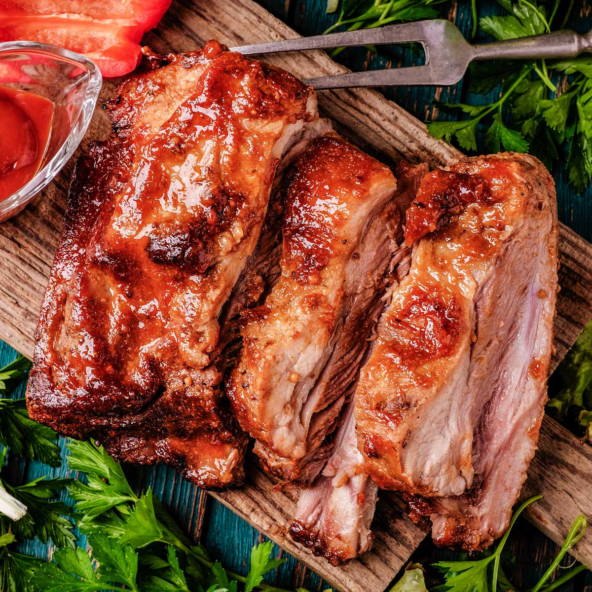 Grilled ribs on a wooden board with garnishes and a small bowl of sauce.