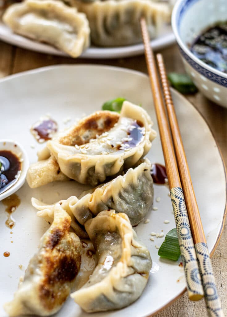 Pan-fried kale veggie dumplings with golden-brown seared bottoms served on a white plate, accompanied by chopsticks, a dipping sauce, and green onions, with more dumplings visible in the background.