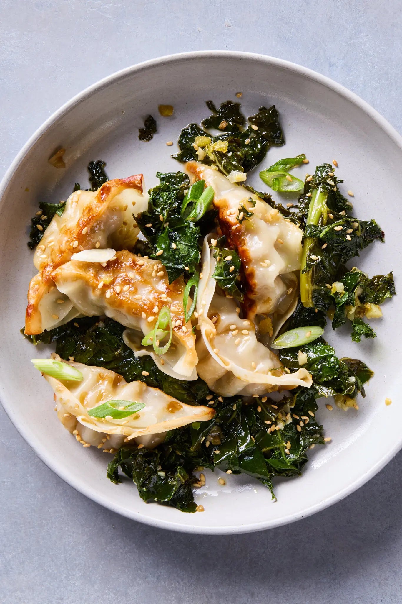 Pan-fried dumplings with golden-brown seared edges, served over dark green cooked kale with chopped green onions and sesame seeds, presented on a light gray plate against a gray background.