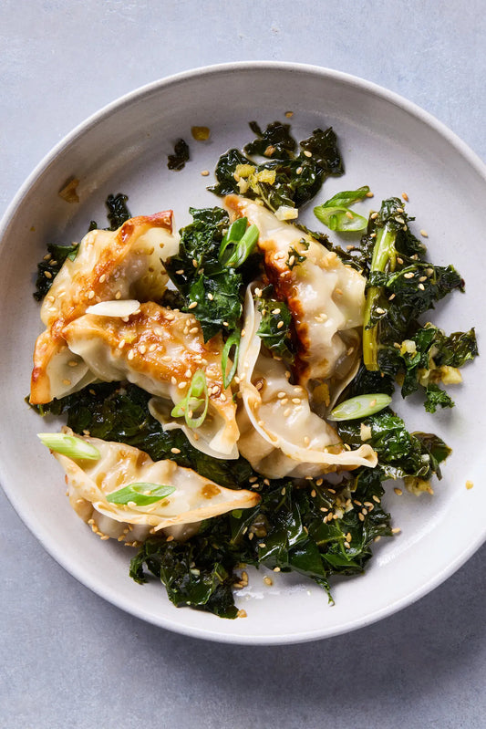 Pan-fried dumplings with golden-brown seared edges, served over dark green cooked kale with chopped green onions and sesame seeds, presented on a light gray plate against a gray background.
